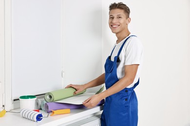 Photo of Smiling handyman with wallpapers and paint roller at window sill indoors