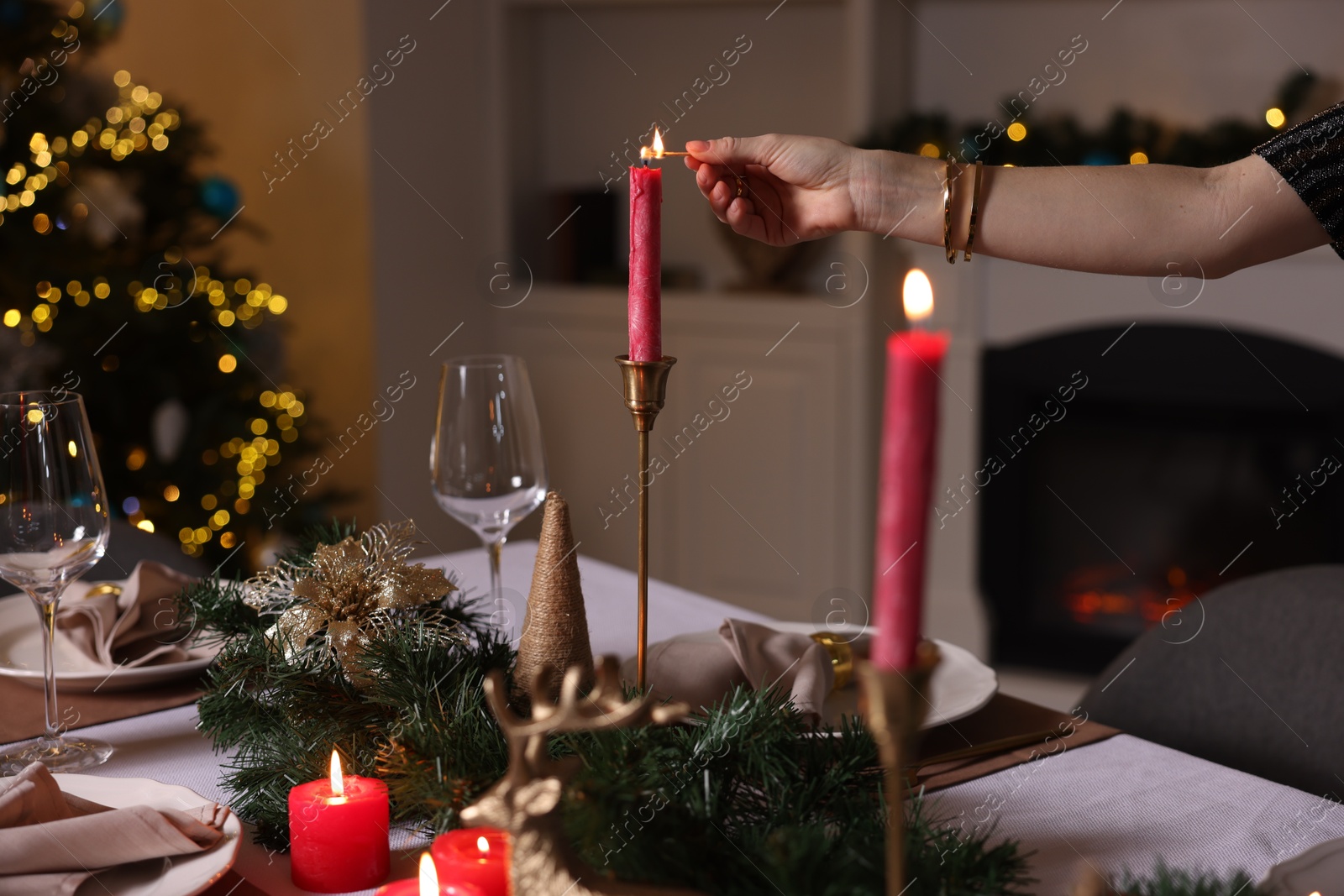 Photo of Woman lighting candle with match at table in room decorated for Christmas, closeup