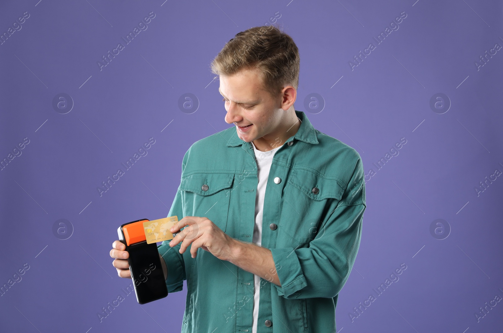 Photo of Happy young man with payment terminal and debit card on purple background