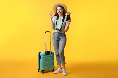 Woman with tickets, passport, suitcase and vintage camera showing ok gesture on orange background