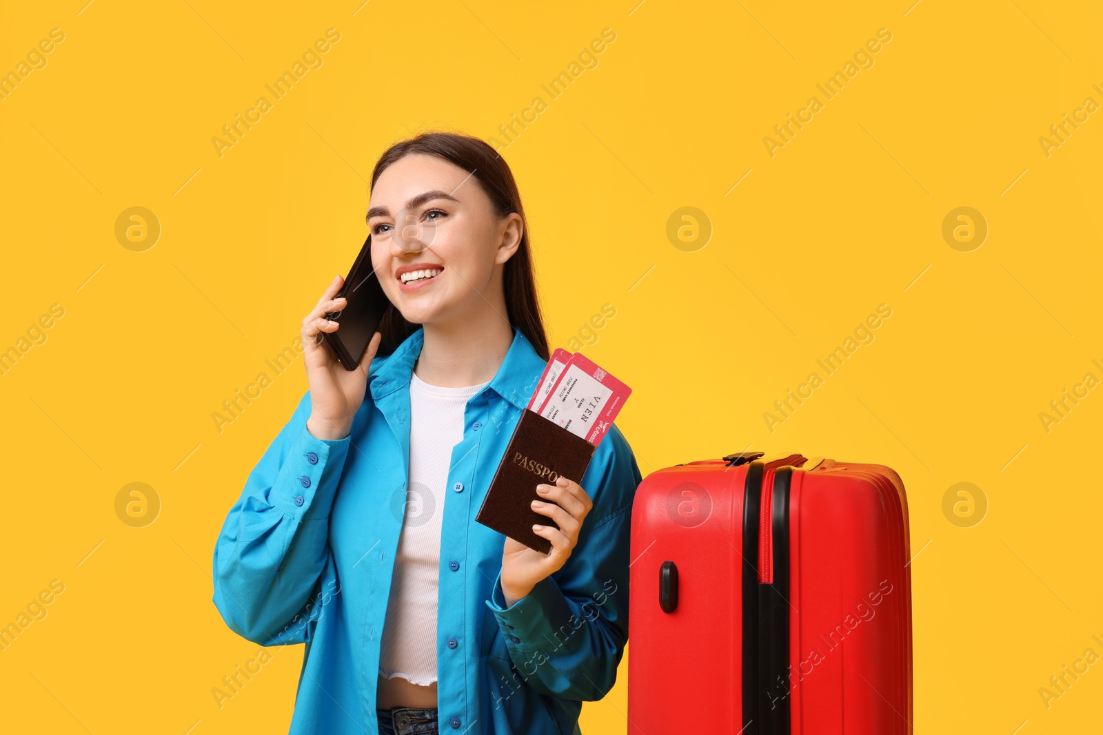 Photo of Happy woman with tickets, passport and suitcase talking on smartphone against orange background