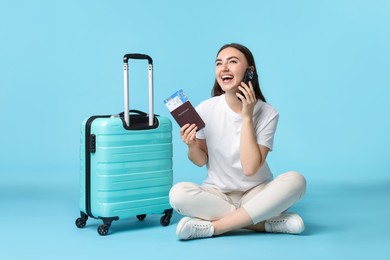 Woman with ticket, passport and suitcase talking on smartphone against light blue background