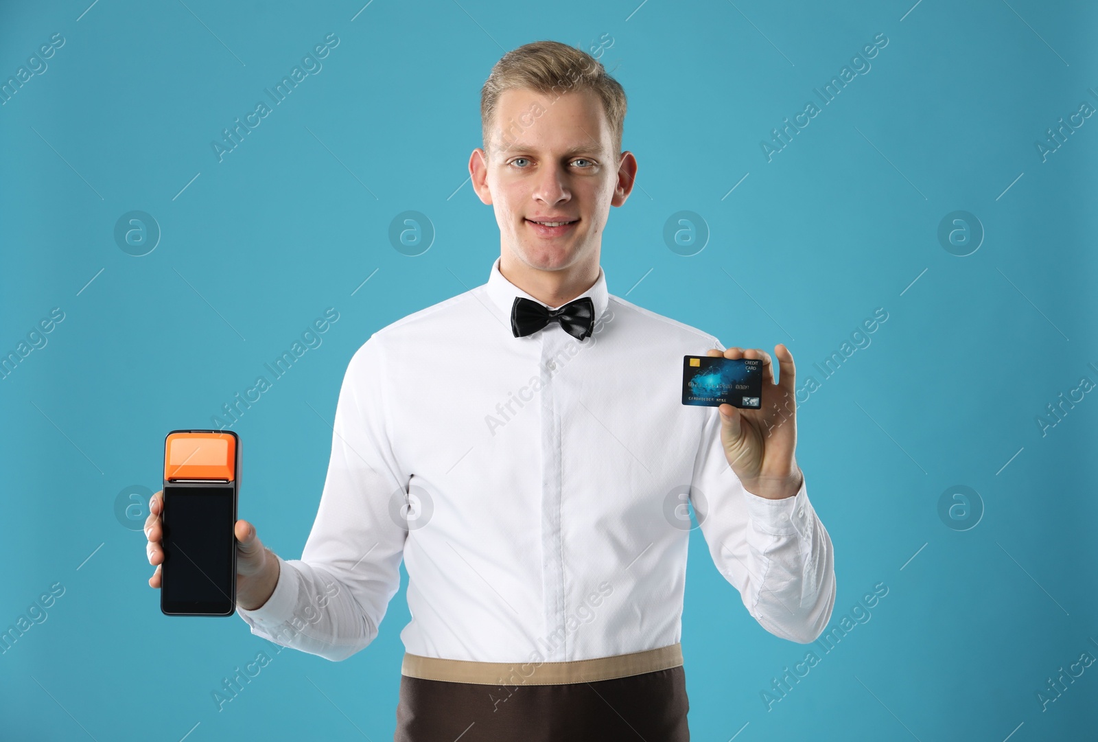 Photo of Happy waiter with payment terminal and debit card on light blue background