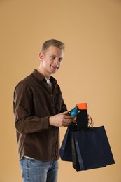 Photo of Happy young man with payment terminal, shopping bags and debit card on beige background