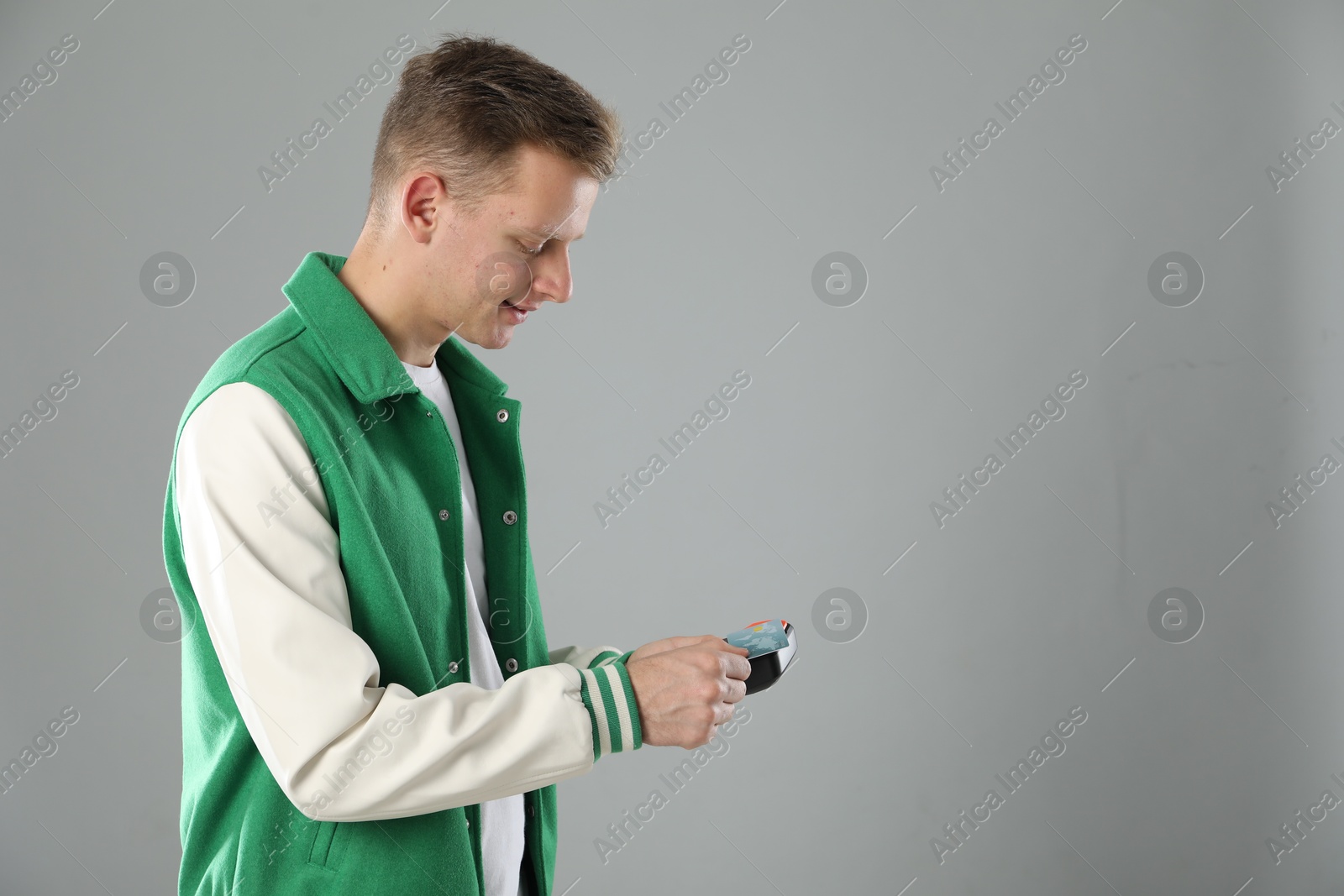 Photo of Happy young man with payment terminal and debit card on light grey background, space for text