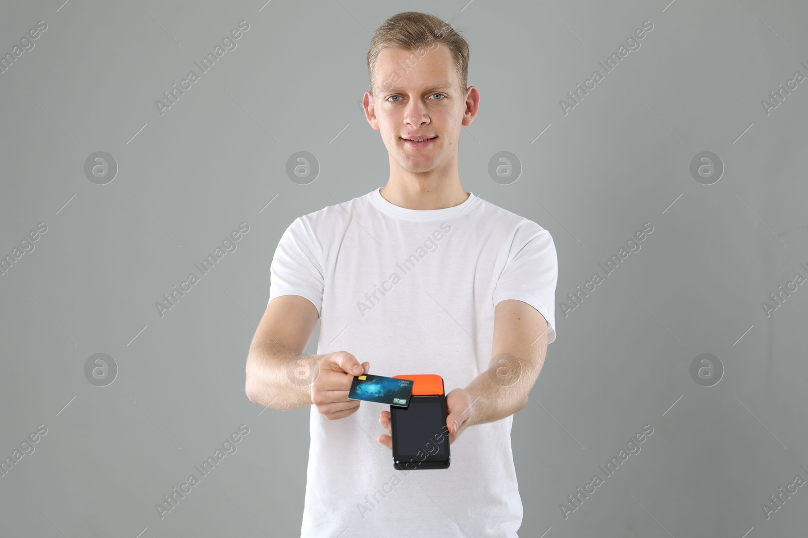 Photo of Happy young man with payment terminal and debit card on light grey background