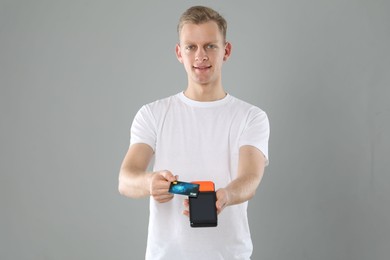 Happy young man with payment terminal and debit card on light grey background