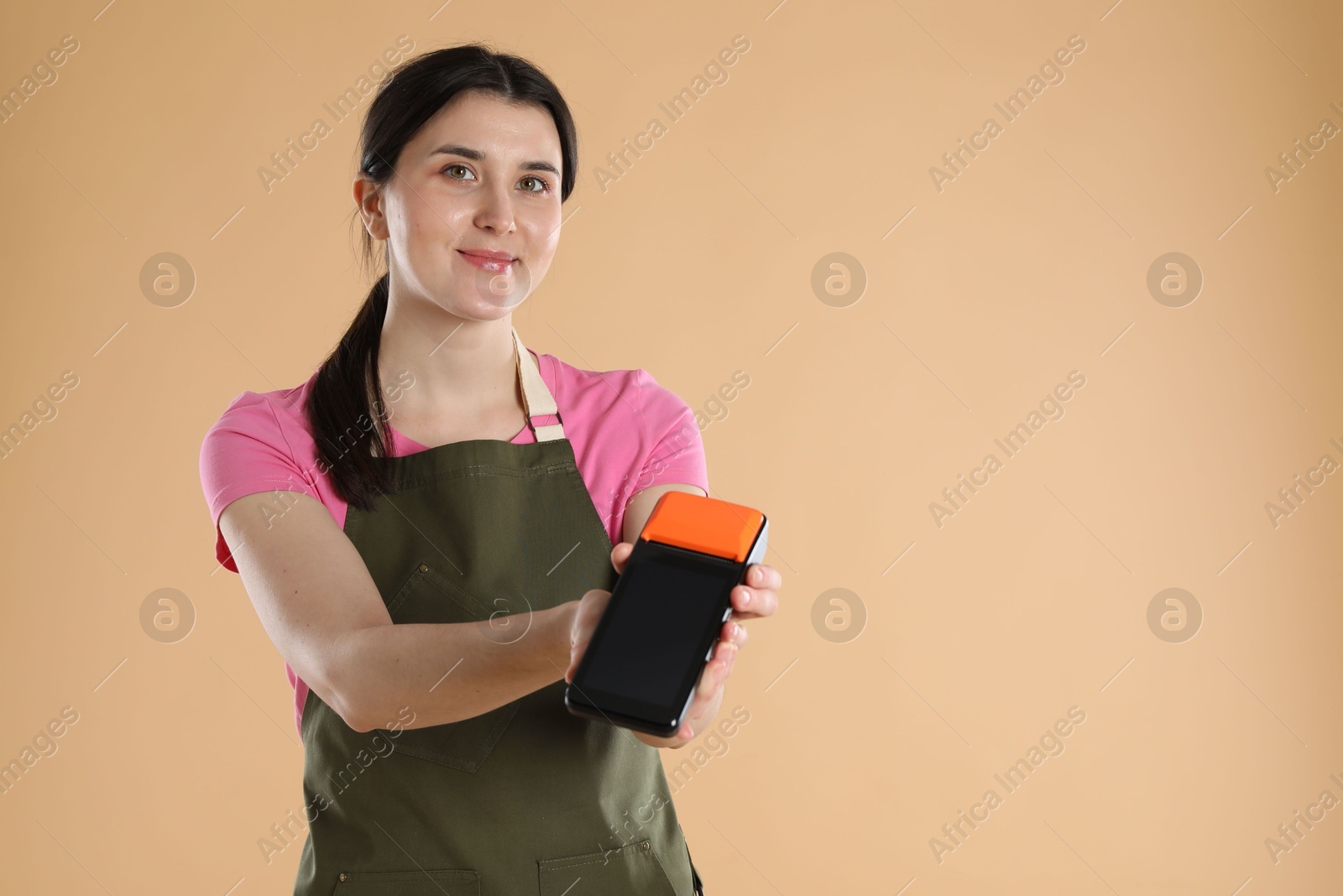 Photo of Young woman in apron with payment terminal on beige background, space for text