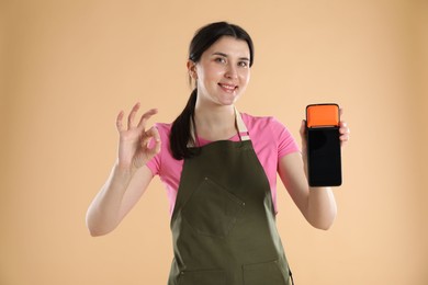 Photo of Happy young woman in apron with payment terminal showing OK gesture on beige background