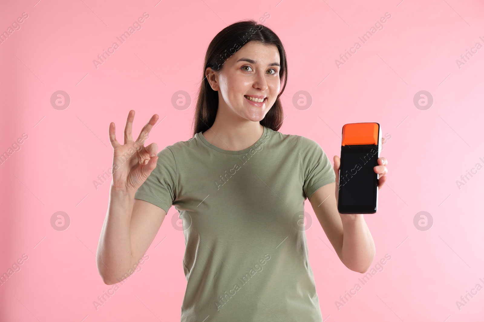 Photo of Happy young woman with payment terminal showing OK gesture on pink background