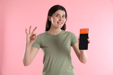 Happy young woman with payment terminal showing OK gesture on pink background