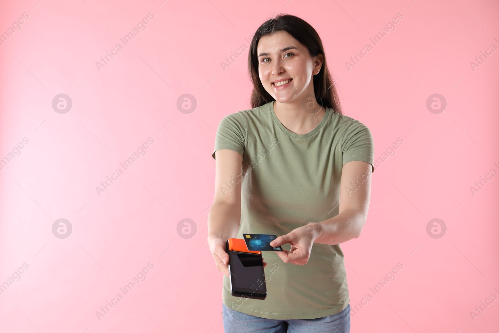 Photo of Happy young woman with payment terminal and debit card on pink background, space for text