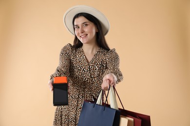 Happy young woman with payment terminal and shopping bags on beige background