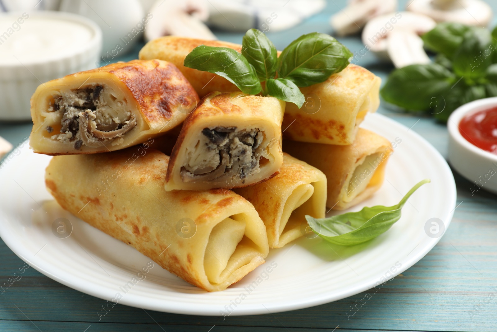 Photo of Delicious rolled crepes with mushrooms and basil on light blue wooden table, closeup