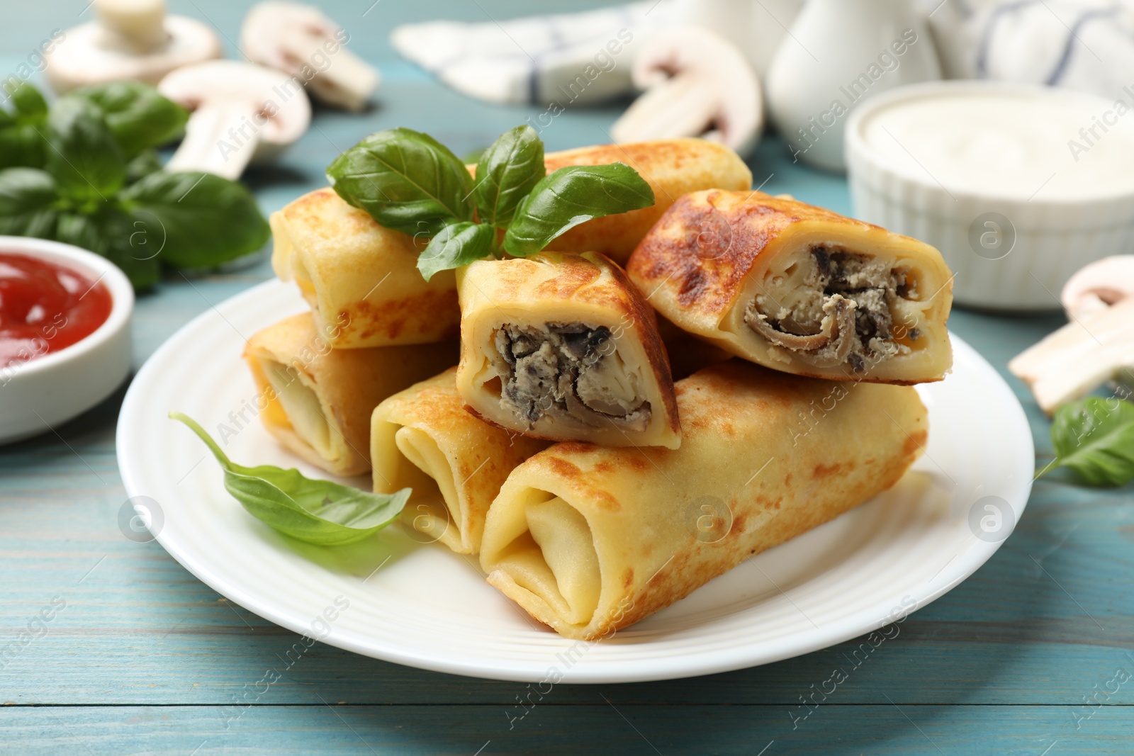 Photo of Delicious rolled crepes with mushrooms and basil on light blue wooden table, closeup