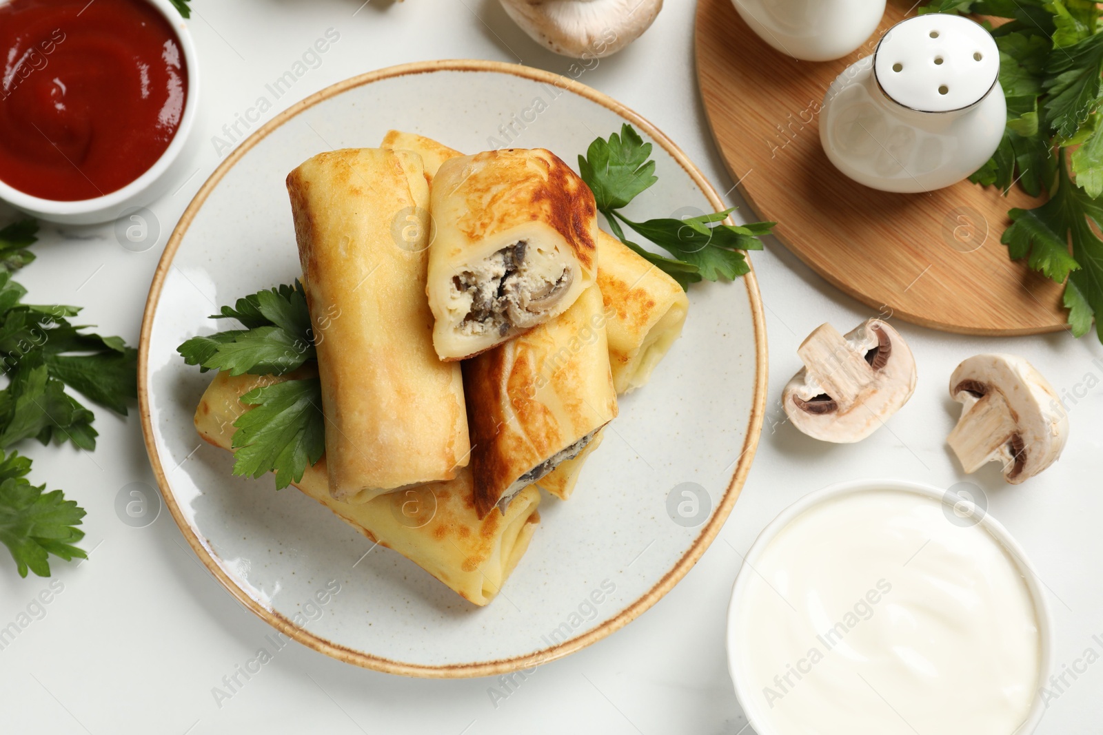 Photo of Delicious rolled crepes with mushrooms, parsley, sauce and sour cream on white marble table, flat lay