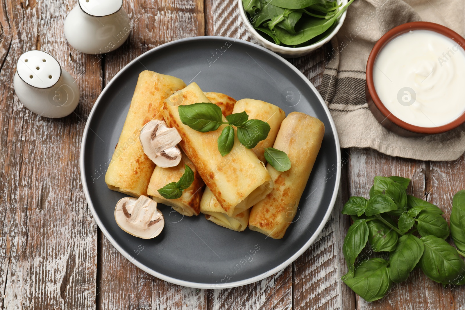 Photo of Delicious rolled crepes with mushrooms, basil and sour cream on wooden table, flat lay