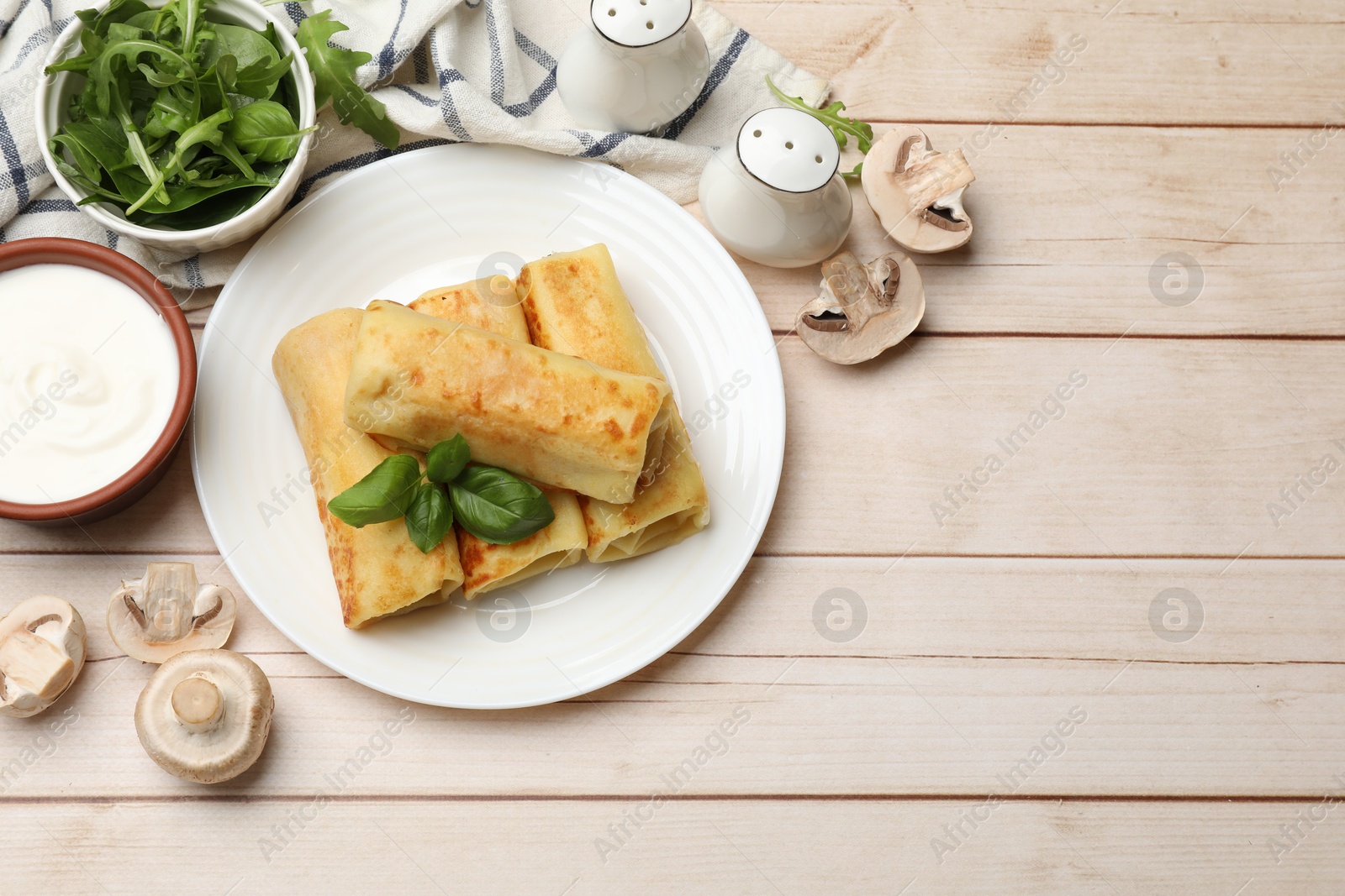 Photo of Delicious rolled crepes with basil, arugula, mushrooms and sour cream on wooden table, flat lay. Space for text