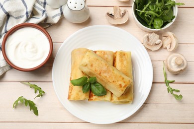 Photo of Delicious rolled crepes with basil, arugula, mushrooms and sour cream on wooden table, flat lay