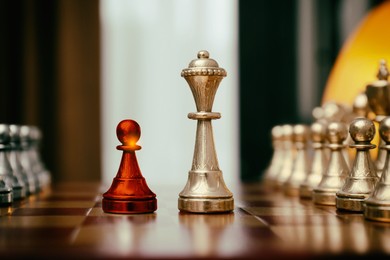 Image of Red pawn standing in front of golden queen on wooden chessboard indoors