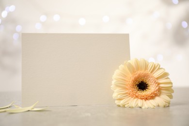 Photo of Blank greeting card and herbera flower on table against blurred festive lights, space for text. Happy Mother's day