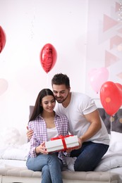 Photo of Young man presenting gift to his girlfriend in bedroom decorated with heart shaped balloons. Valentine's day celebration