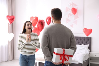 Photo of Young man presenting gift to his girlfriend in bedroom decorated with heart shaped balloons. Valentine's day celebration