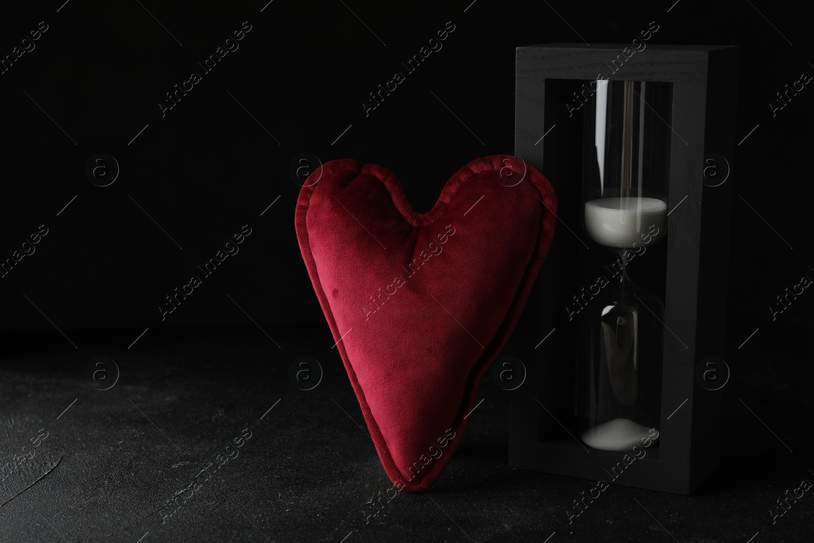 Photo of Heart shaped cushion and hourglass on black stone table, space for text. Relationship problems concept