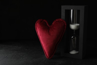 Photo of Heart shaped cushion and hourglass on black stone table, space for text. Relationship problems concept