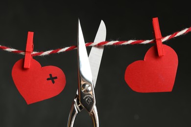 Photo of Red paper hearts on rope and scissors against black background. Composition symbolizing problems in relationship