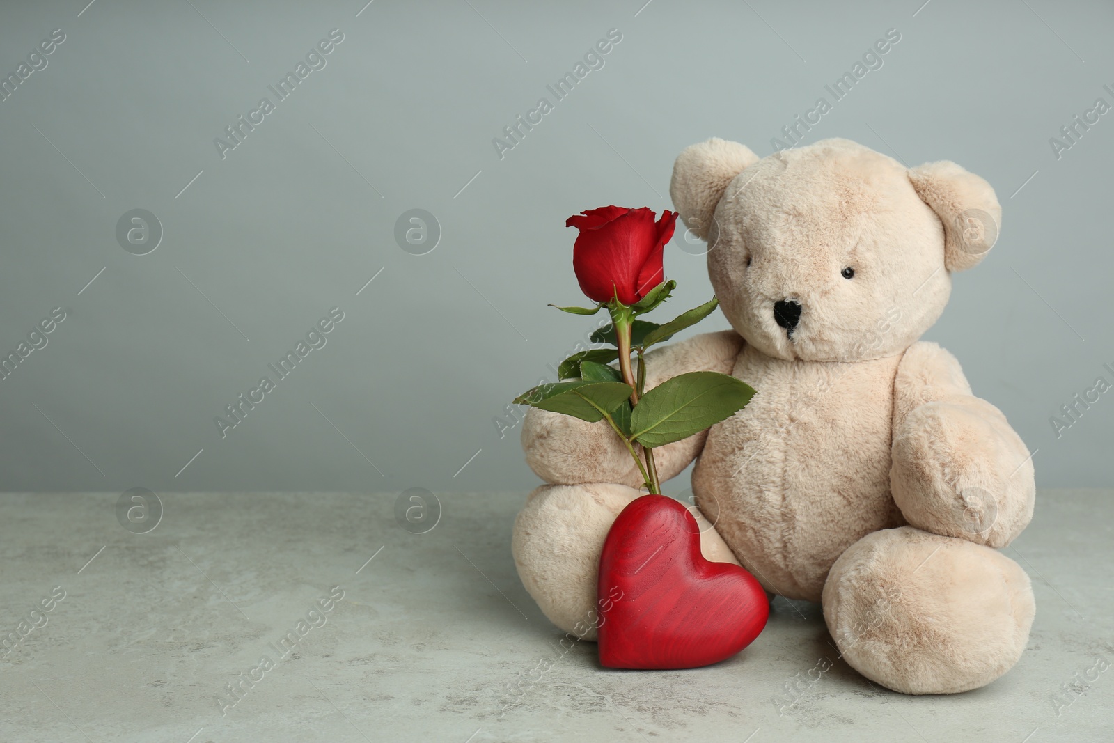 Photo of Cute teddy bear with heart and red rose on light grey stone table, space for text. Valentine's day celebration