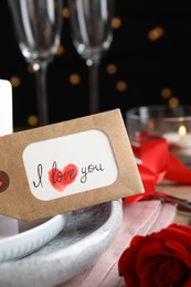 Photo of Beautiful place setting and tag with phrase I Love You on table against blurred lights, closeup. Valentine's day romantic dinner