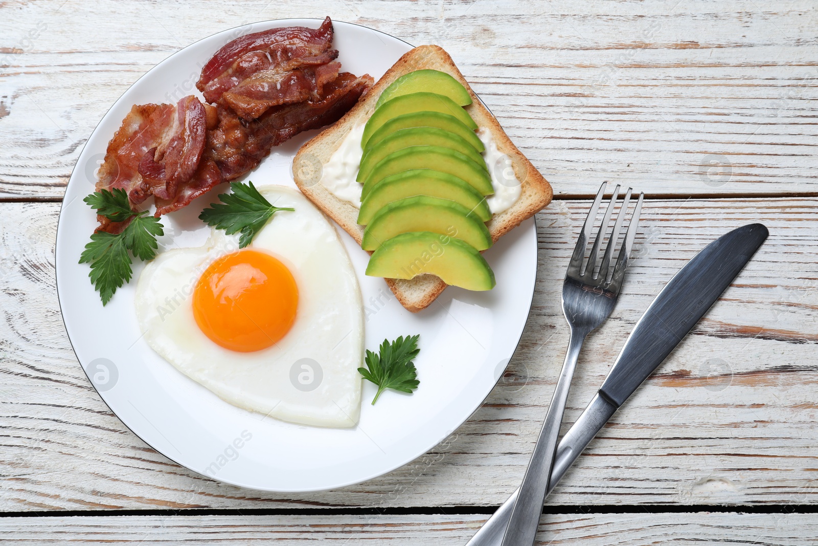 Photo of Romantic breakfast with fried bacon, heart shaped egg and avocado toast on white wooden table, flat lay. Valentine's day celebration