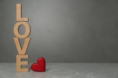 Photo of Word Love made of wooden letters near red decorative heart on light grey table. Space for text