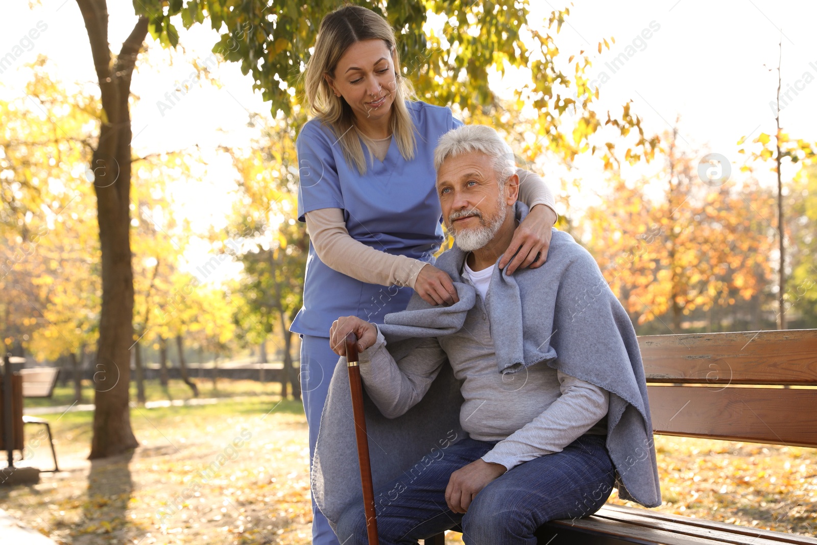 Photo of Caregiver assisting senior man on wooden bench in park. Home health care service