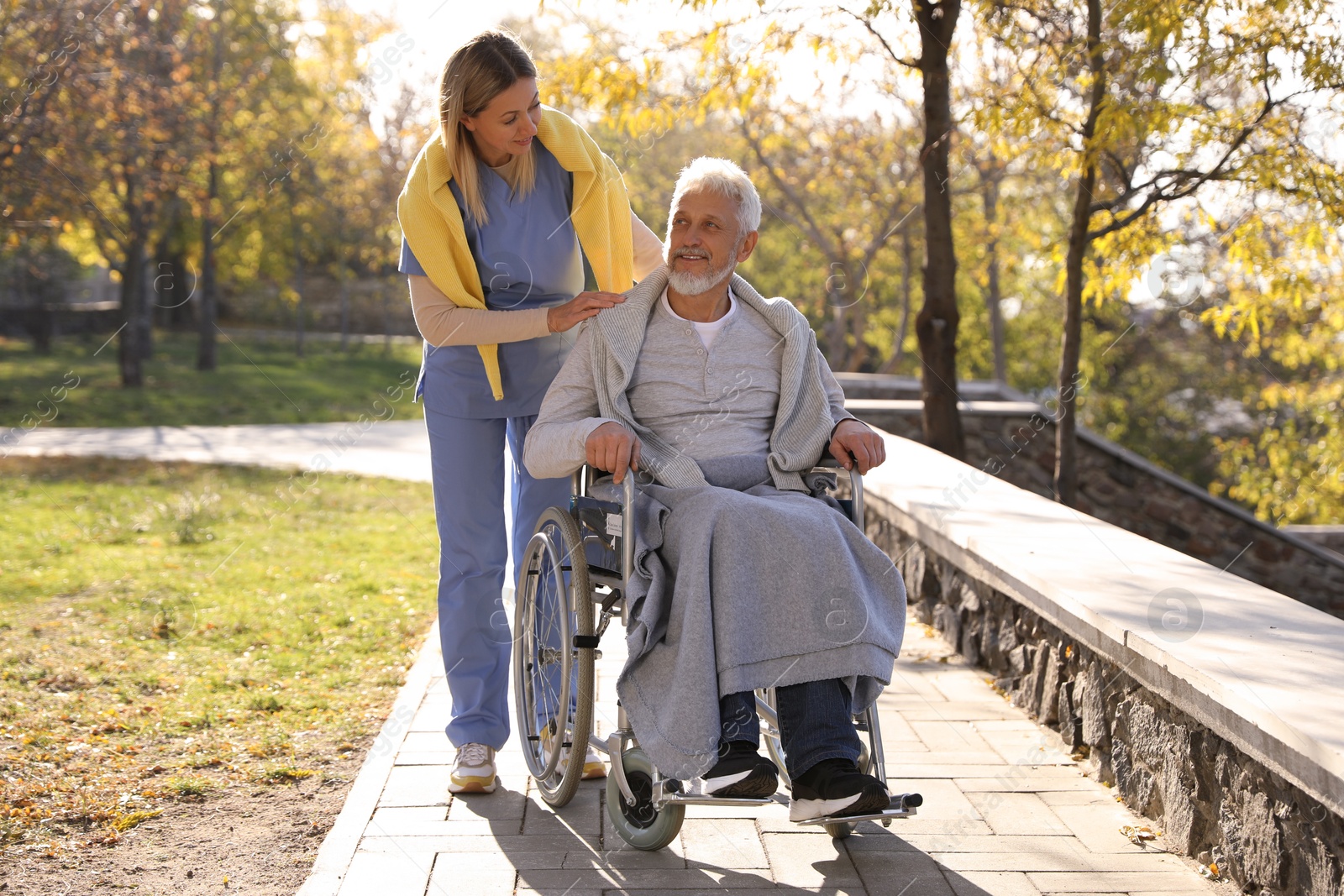 Photo of Caregiver assisting senior man on wheelchair in park. Home health care service