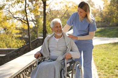 Photo of Caregiver assisting senior man on wheelchair in park. Home health care service