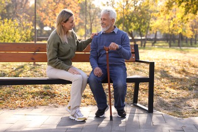 Photo of Caregiver assisting senior man on wooden bench in park. Home health care service