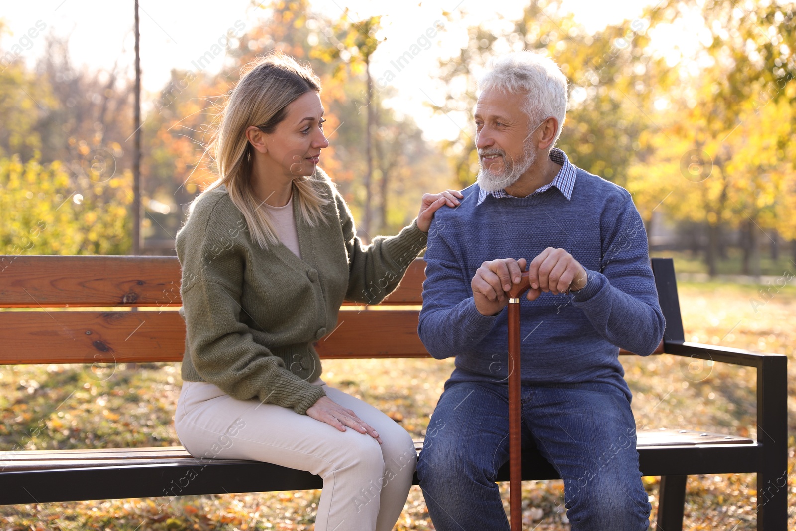 Photo of Caregiver assisting senior man on wooden bench in park. Home health care service