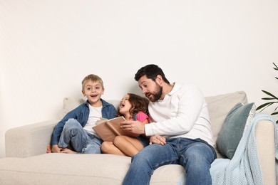 Overwhelmed father reading book to his happy children on sofa at home