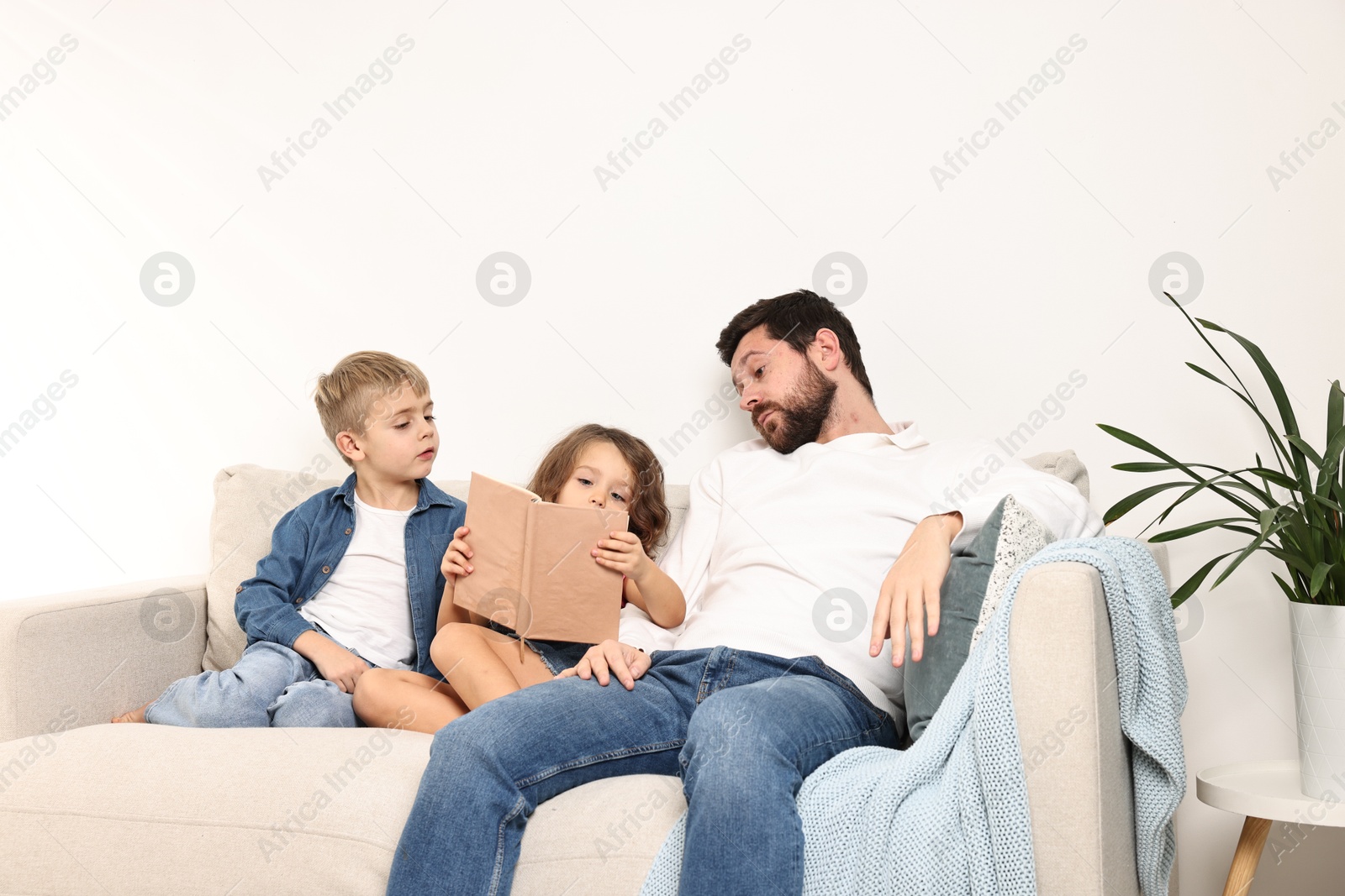 Photo of Overwhelmed father and his children reading book on sofa at home