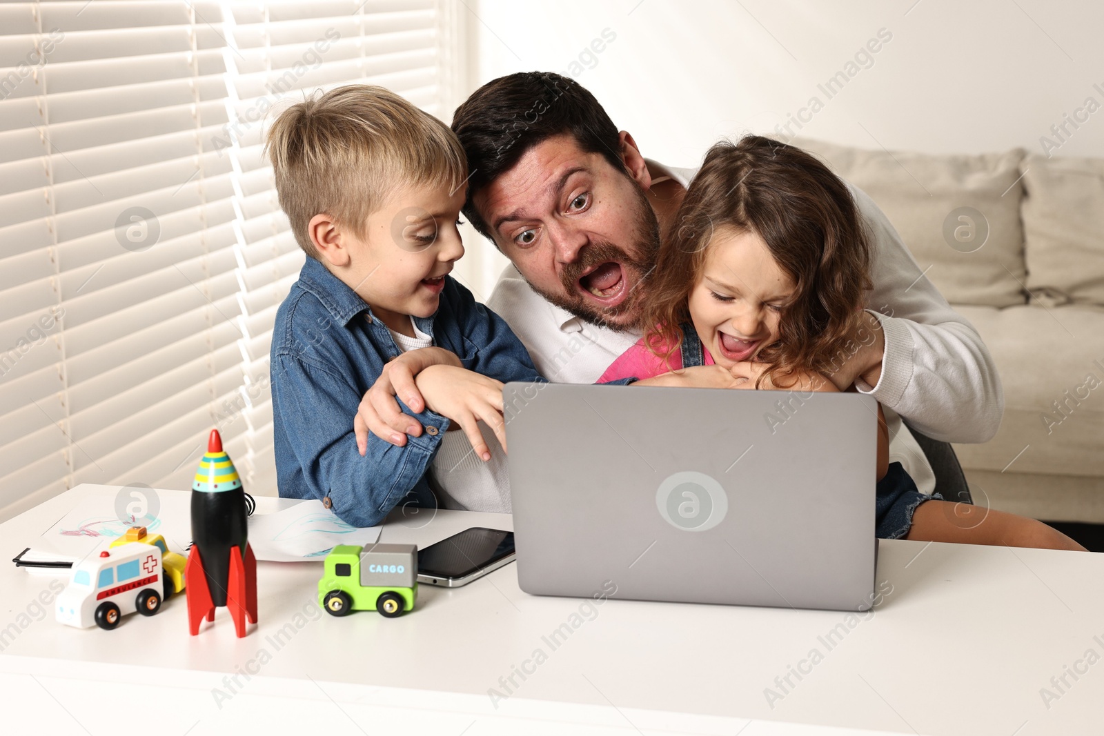 Photo of Naughty children disturbing their overwhelmed father at table with laptop indoors