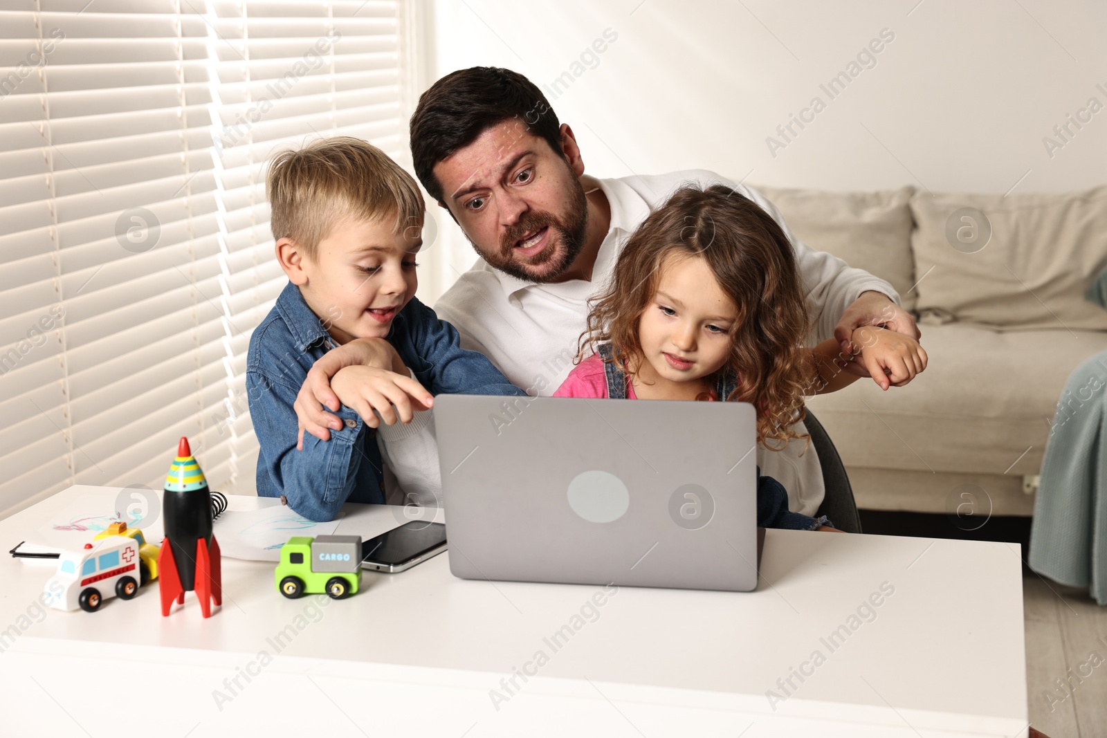 Photo of Naughty children disturbing their overwhelmed father at table with laptop indoors