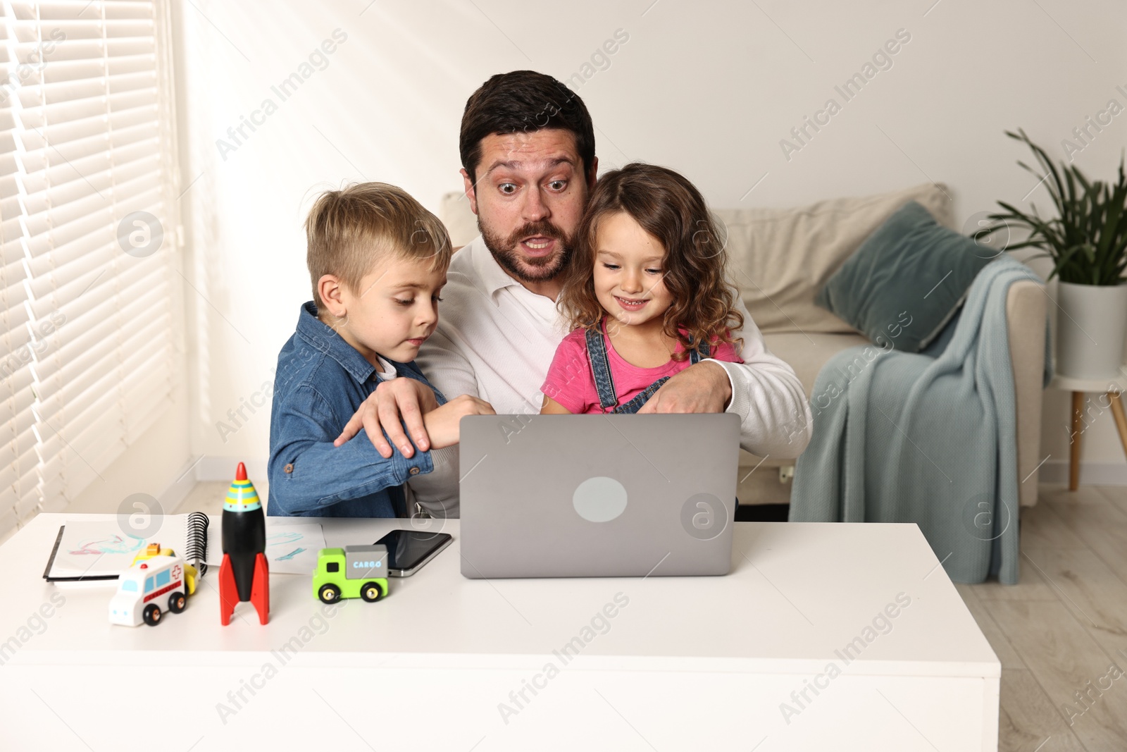 Photo of Naughty children disturbing their overwhelmed father at table with laptop indoors