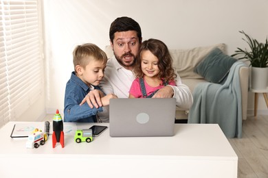 Photo of Naughty children disturbing their overwhelmed father at table with laptop indoors