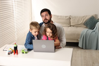 Photo of Naughty children disturbing their overwhelmed father at table with laptop indoors