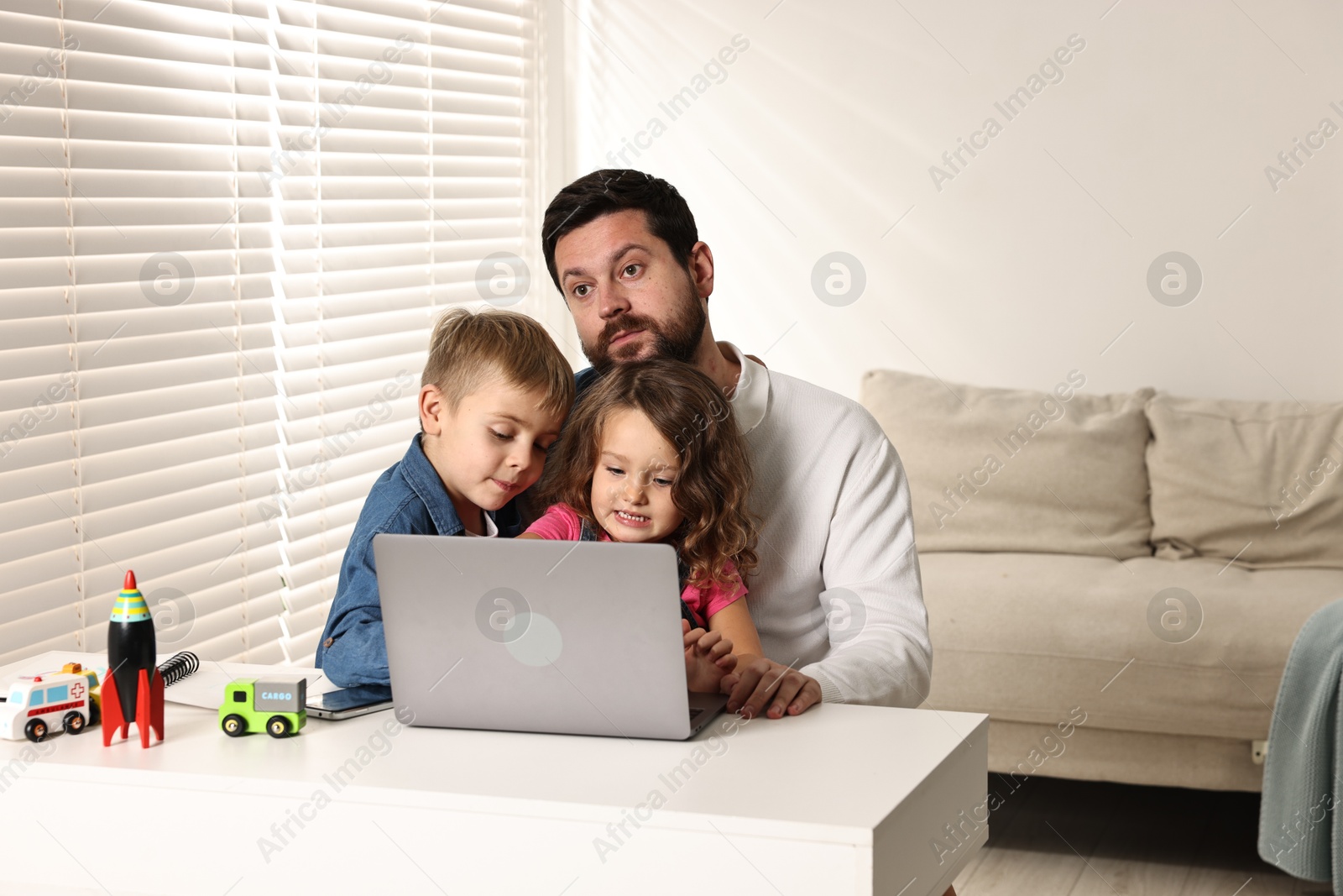 Photo of Naughty children disturbing their overwhelmed father at table with laptop indoors