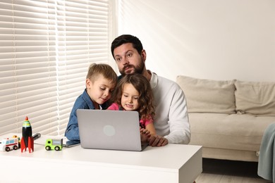 Photo of Naughty children disturbing their overwhelmed father at table with laptop indoors