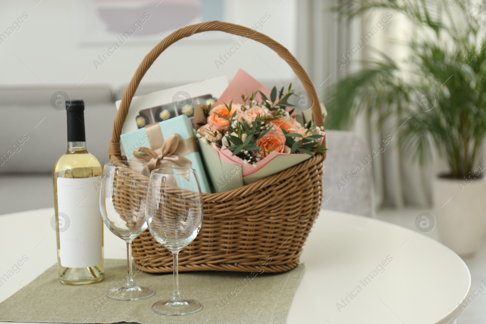 Photo of Wicker basket with gifts near bottle of wine and glasses on table indoors