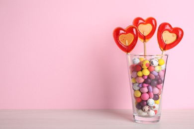 Photo of Delicious heart shaped lollipops and dragees in glass on table against pink background. Space for text